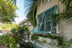 View of a home with light blue Bahama shutters surrounded by greenery